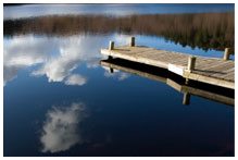 A pier amongst some reeds in clear water with the sky reflecting on it